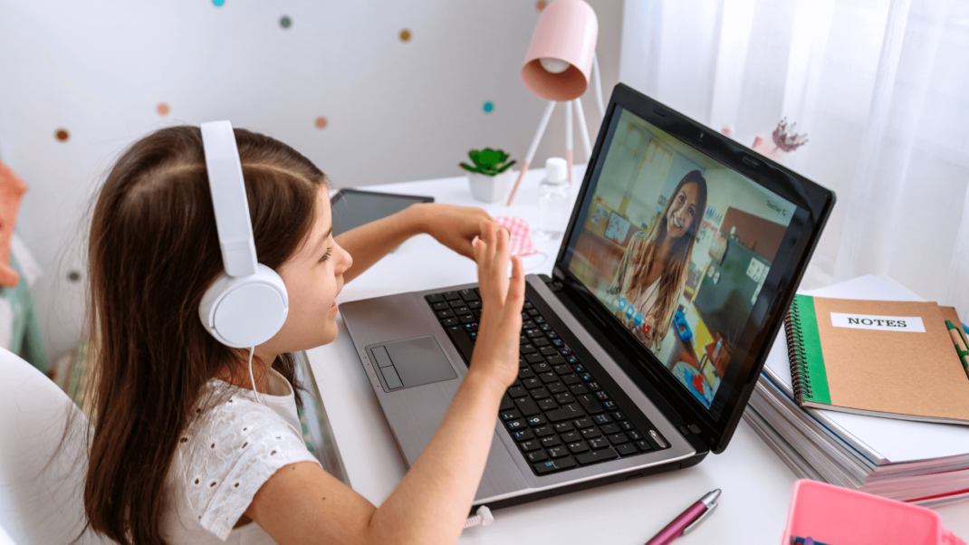 A young girl wearing headphones sits at a desk, waving at a woman on a video call on her laptop, with notebooks and stationery nearby.