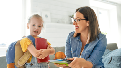 A woman hands a young girl with a backpack a stack of books and an apple. The girl is smiling and holding a red notebook.