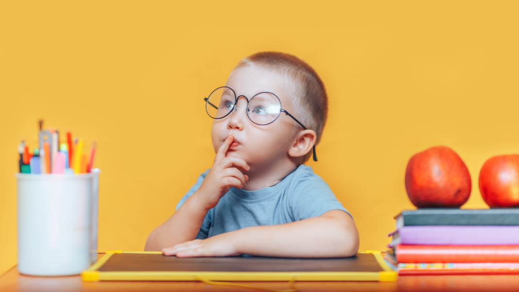 A young child wearing glasses sits at a desk with a blackboard, colored pencils, apples, and books, holding a finger to their lips in a thoughtful pose against a yellow background.