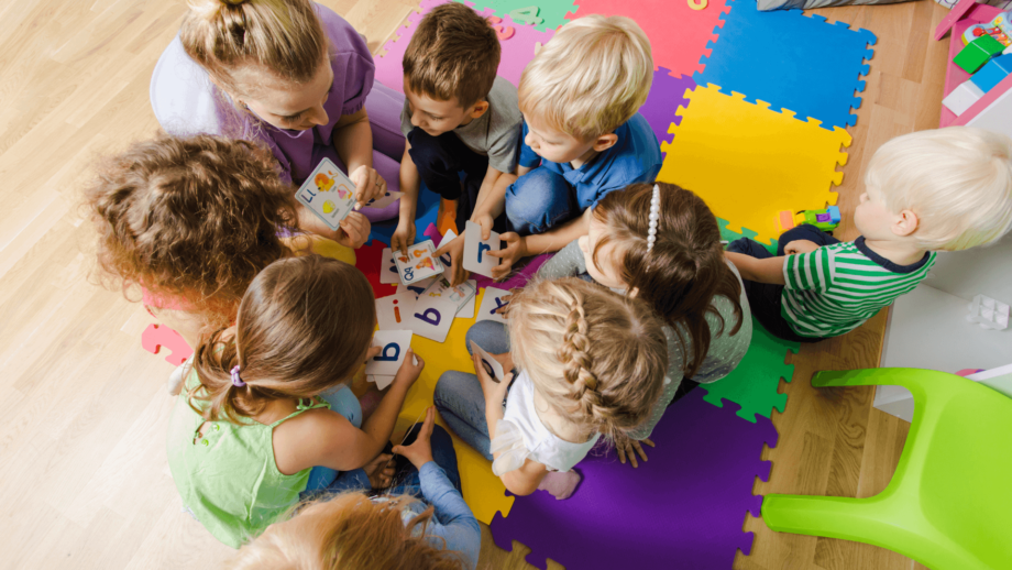 A group of young children sit in a circle on colorful foam mats, learning letters with the help of an adult.