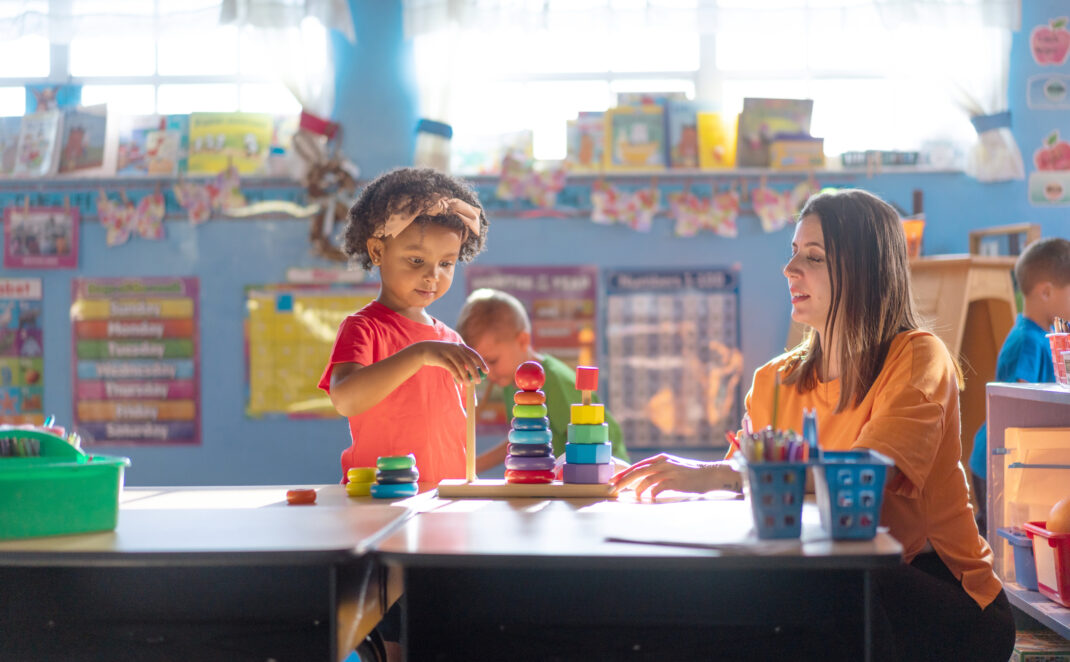 A young girl stacks colorful toys while a woman, likely a teacher, sits nearby at a table in a brightly lit classroom.