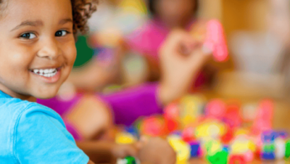A young child smiles at the camera while sitting at a table, playing with colorful plastic building blocks. Other children are visible in the blurred background.
