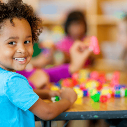 A young child smiles at the camera while sitting at a table, playing with colorful plastic building blocks. Other children are visible in the blurred background.