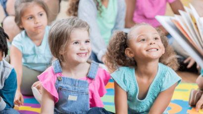 A group of young children sit on a colorful mat, smiling and looking toward an adult who is reading a book to them.