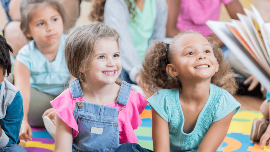 A group of young children sit on a colorful mat, smiling and looking toward an adult who is reading a book to them.