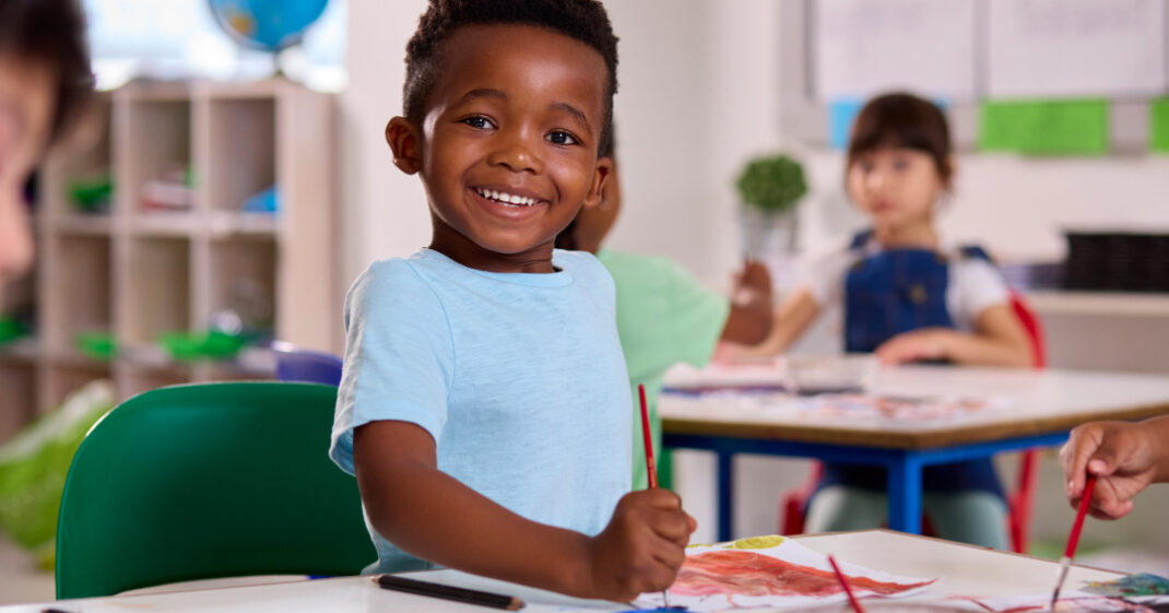 Smiling child in a light blue shirt holds a paintbrush and paints at a table in a classroom, with other children working in the background.